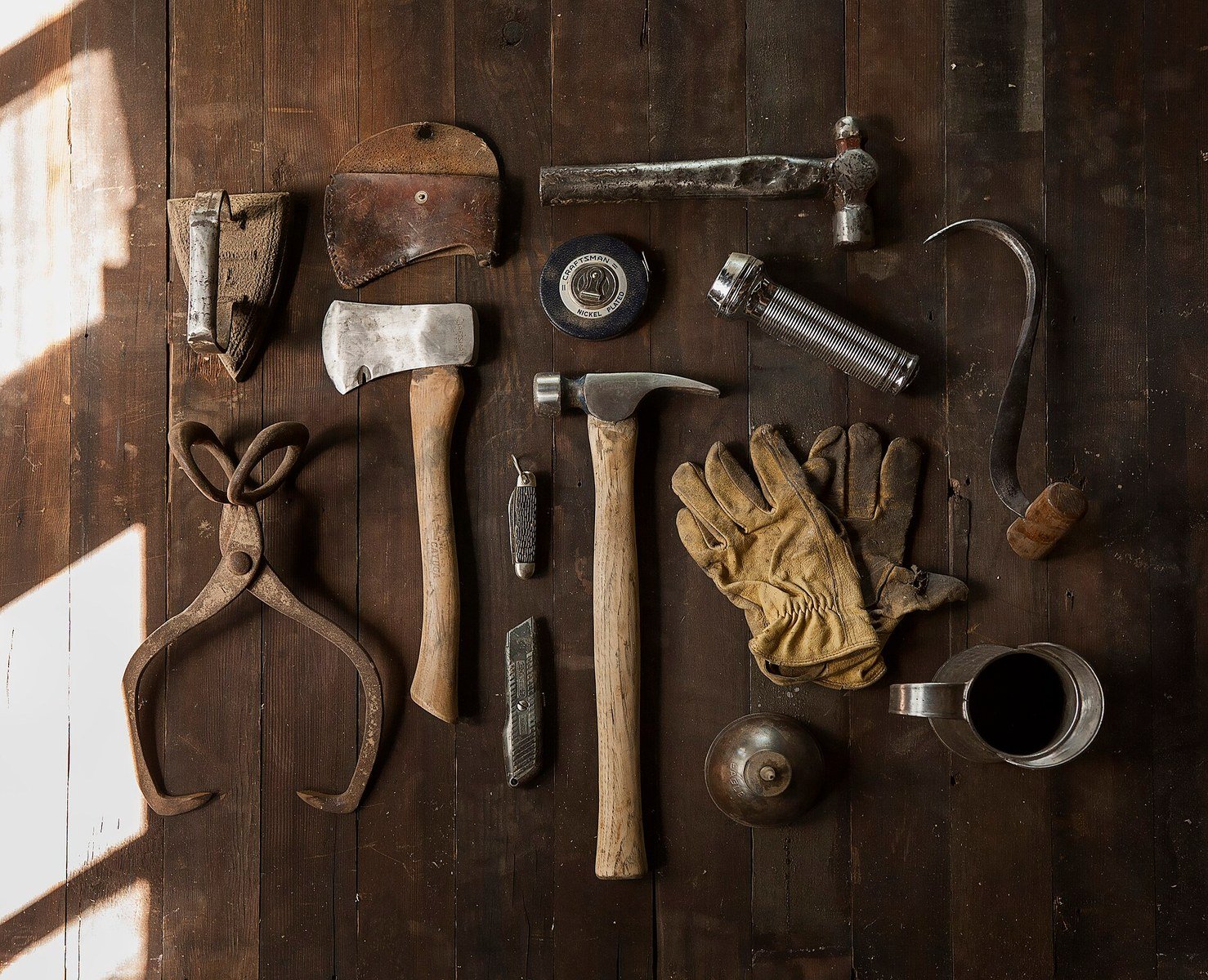 A flat-lay arrangement of carpenter and construction hand tools on a dark wooden surface, including an axe, claw hammer, tape measure, pliers, utility knife, work gloves, torch, and other assorted tools