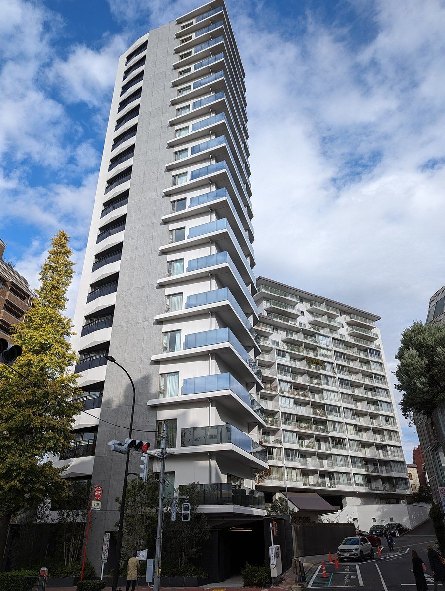 A tall modern white residential high-rise building with glass balconies on every floor, viewed from street level, with traffic lights and trees in the foreground under a partly cloudy sky