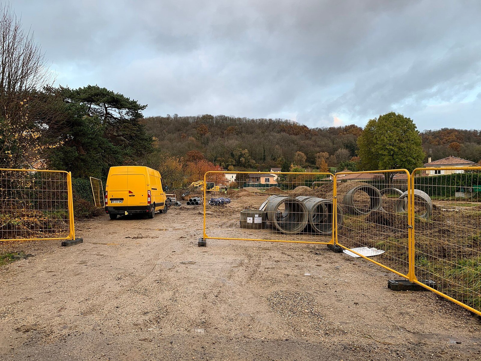 An outdoor construction site with yellow temporary security fencing at the entrance, a yellow van parked on a dirt ground, large concrete pipe sections and construction materials stacked nearby, surrounded by trees with autumn foliage under a cloudy sky