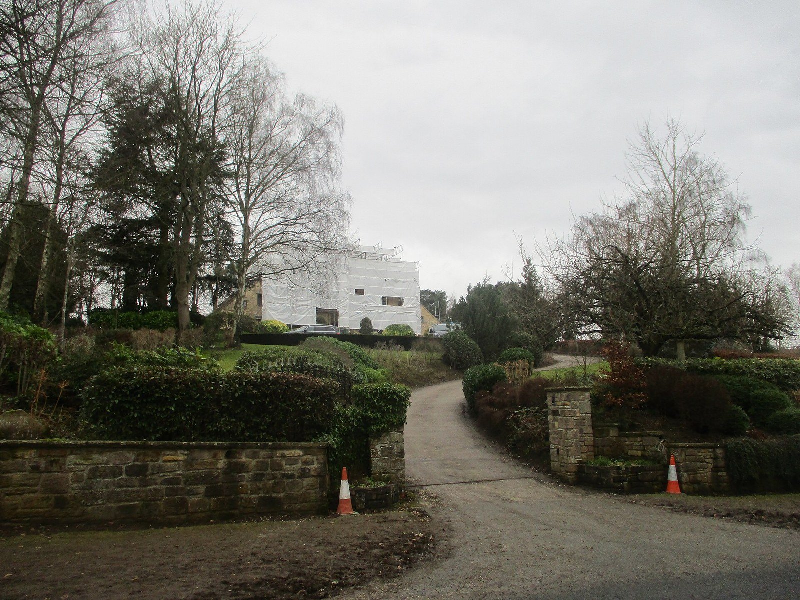 A house undergoing renovation, fully wrapped in white protective scaffolding sheeting, visible from the entrance driveway flanked by stone pillars and orange traffic cones, surrounded by leafless winter trees and garden shrubs in West Yorkshire, England.