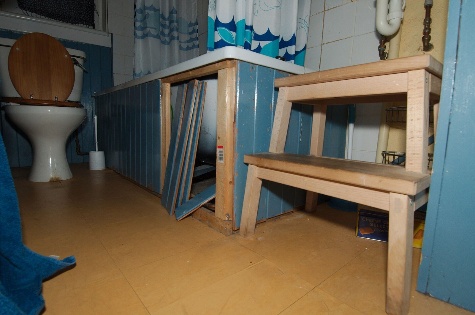 A bathroom undergoing renovation, showing an exposed bathtub frame with blue panelling partially removed, a wooden step stool, tiled flooring, a white toilet, and blue-painted walls with old tiles